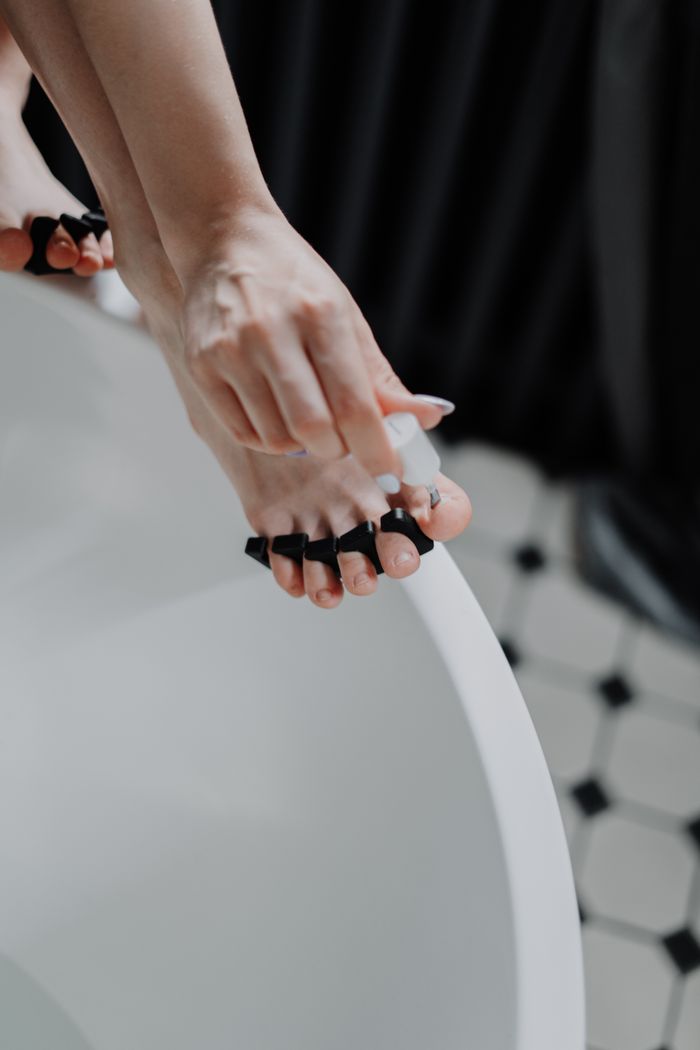 Woman applying nail polish with toe separators during a pedicure session in a ceramic bathtub.